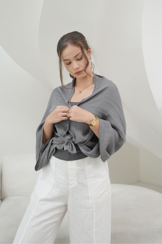 A woman indoors adjusts her Olive Basics Multi-functional Nursing Swaddle, styled as a nursing cover over a black top and white pants.