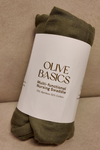An Olive Basics Multi-functional Nursing Swaddle in olive, made from breathable bamboo cotton, is rolled up and placed on a beige surface.