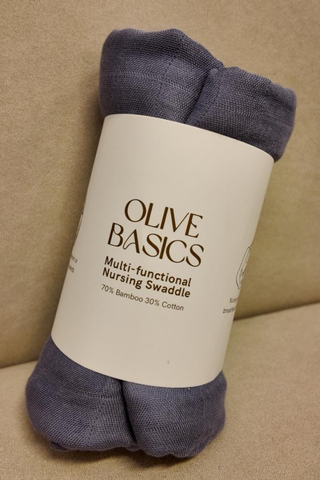 An Ash Blue Olive Basics Multi-functional Nursing Swaddle, made of 70% bamboo and 30% cotton, is rolled with a label on a beige surface. This breathable baby blanket also serves as a convenient nursing cover.