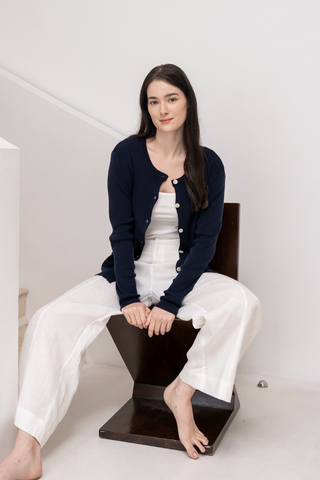 A woman with long dark hair sits barefoot on a chair, wearing the Olive Basics Ellie Reversible Cardigan in TENCEL™ Knit over a white top and loose white pants, posed against a plain white background.