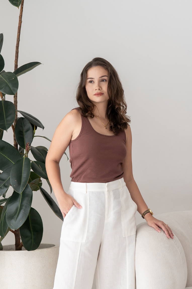A woman with long brown hair wears the brown Esther Easy Latch Tank by Olive Basics and white trousers, standing indoors near a plant with one hand resting on a white chair.