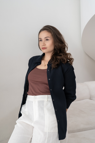 A woman with long brown hair, wearing a black shirt over the Olive Basics Esther Easy Latch Tank and white pants, sits on a light-colored sofa against a plain white wall.