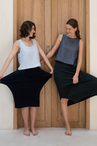 Two women stand barefoot before wooden doors, each in a Leah Super Stretch Slit Skirt by Olive Basics, holding out the sides to show the stretchy fabric’s width while wearing sleeveless tops.