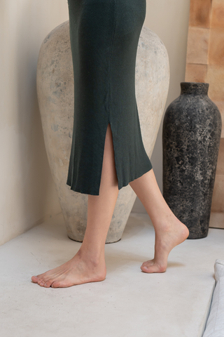 A person stands barefoot on a light floor in front of two large vases, wearing the Olive Basics Leah Super Stretch Slit Skirt in dark green.