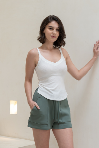 A woman stands indoors against a plain light wall, wearing Olive Basics' Naomi Stretchy Pocket Shorts in khaki green with one hand in her pocket and the other resting on the wall.