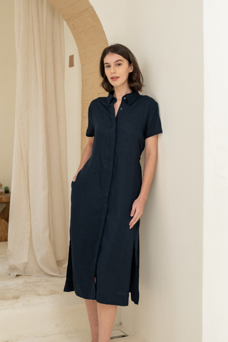 A woman stands indoors against a light wall, wearing the Olive Basics Marie Short Sleeve Midi Dress in navy TENCEL linen blend, with front buttons and pockets.