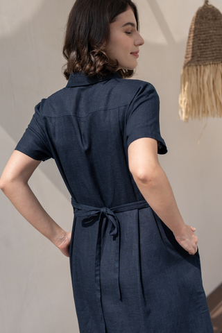 A woman stands indoors with her back to the camera, wearing the Olive Basics Marie Short Sleeve Midi Dress in navy blue TENCEL linen blend, tied at the waist, near a hanging woven decoration.