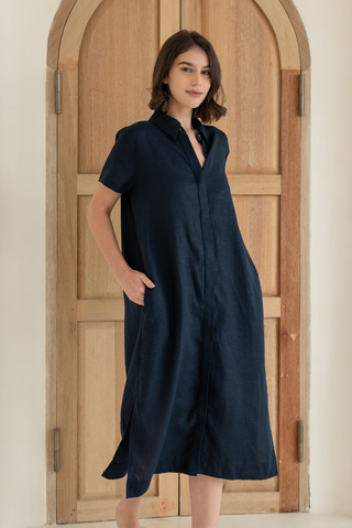 A woman stands barefoot indoors in front of a wooden arched door, wearing the Olive Basics Marie Short Sleeve Midi Dress—a loose, navy blue TENCEL linen blend midi dress with short sleeves and pockets.