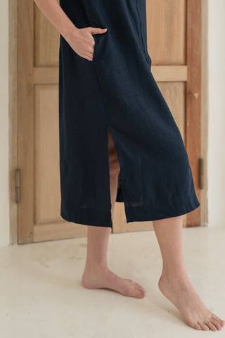 A person stands barefoot on a light floor before a wooden door, wearing the Olive Basics Marie Short Sleeve Midi Dress in navy blue, made from a TENCEL linen blend with short sleeves and a side slit.