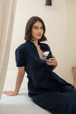 A woman wearing the Olive Basics Marie Short Sleeve Midi Dress sits on a bed, holding a white electronic device to her chest and looking at the camera—ideal for easy breastfeeding access.