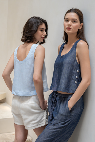 Two women pose indoors: one wears a light blue TENCEL linen sleeveless top with white shorts, while the other sports the Olive Basics Leigh-Anne Reversible Button Top in denim blue with matching pants.