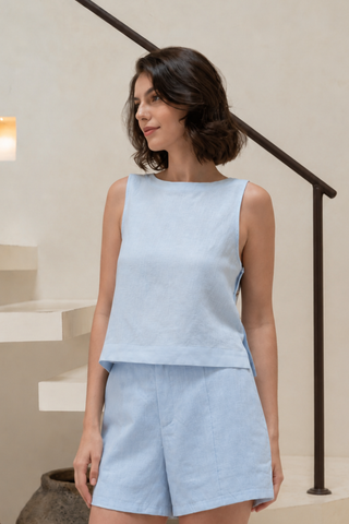A woman stands indoors near a staircase, looking to the side, wearing the Olive Basics Leigh-Anne Reversible Button Top in Sky Blue with matching shorts.