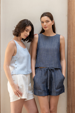 Two women stand side by side: one in a light blue sleeveless TENCEL linen top with white shorts, the other in the Olive Basics Leigh-Anne Reversible Button Top in navy, paired with matching shorts and hands in her pockets.
