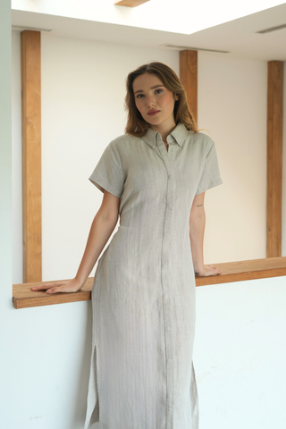 Sure! Here’s a rewritten version using your product data: A woman stands indoors, leaning against a wooden railing, wearing the Olive Basics Marie Short Sleeve Midi Dress—a light-colored button-down crafted from a soft TENCEL™ linen blend. This chic dress features convenient breastfeeding access, perfectly balancing style and practicality.