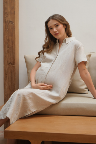 A pregnant woman wearing the Olive Basics Marie Short Sleeve Midi Dress in light beige, made from a TENCEL™ Linen Blend, sits on a cushioned bench, resting her hand on her belly and looking at the camera with a neutral expression.