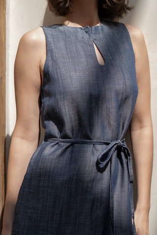 A woman in the Noel Reversible Mini Dress by Olive Basics—a sleeveless, belted blue chambray dress in TENCEL™ linen blend with a keyhole neckline—stands in natural light.