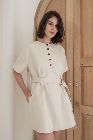 A woman stands indoors in front of a wooden door, wearing the Olive Basics Joanna Reversible T-shirt Pocket Dress in beige with short sleeves, pockets, front buttons, and her hand in her pocket.