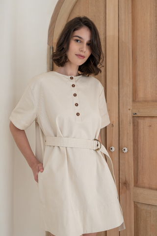 A woman with shoulder-length brown hair wears the Olive Basics Joanna Reversible T-shirt Pocket Dress in beige, standing indoors in front of wooden doors.