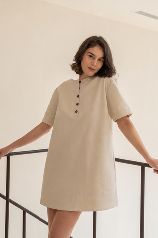 A woman wears the Olive Basics Joanna Reversible T-shirt Pocket Dress in beige, featuring short sleeves, a pocket, and front buttons. She stands indoors holding a black railing and looks at the camera.