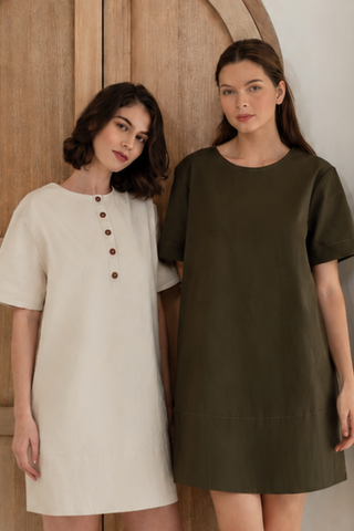 Two women stand indoors—one wears the Joanna Reversible T-shirt Pocket Dress in beige from Olive Basics, while the other wears a loose short-sleeved olive green dress.