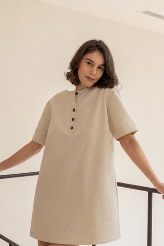 A person with short brown hair stands indoors by a railing, wearing Olive Basics' Joanna Reversible T-shirt Pocket Dress in beige, knee-length TENCEL™ Cotton Canvas with short sleeves and a buttoned placket.