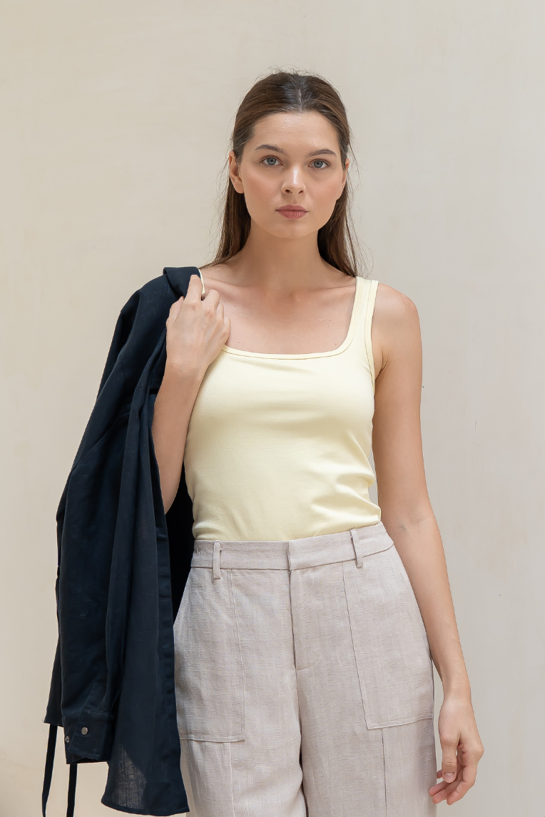 A woman stands against a plain background wearing the Olive Basics Esther Easy Latch Tank in Modal Cotton, beige pants, and holding a black jacket over her shoulder.