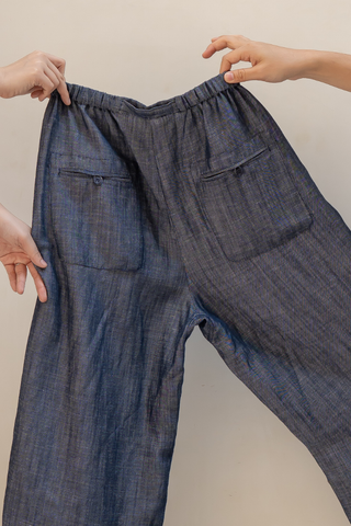 Two hands hold up a pair of Bella Multi-Pocket Drawstring Pants by Olive Basics in dark blue, featuring a TENCEL linen blend, elastic waistband, and multiple pockets, set against a plain background.