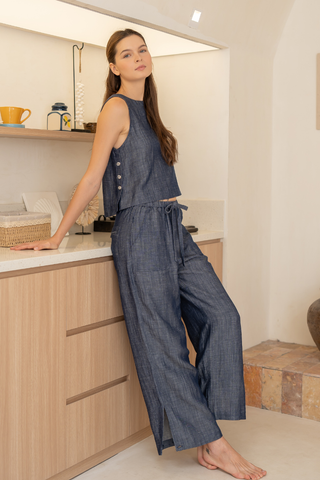 A woman wearing Olive Basics' Bella Multi-Pocket Drawstring Pants, part of a sleeveless blue TENCEL linen blend two-piece set, leans against a kitchen counter in a modern, minimalistic room.