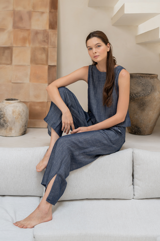 A woman with long brown hair sits barefoot on a light gray sofa, wearing Olive Basics’ Bella Multi-Pocket Drawstring Pants, with neutral décor and pottery in the background.