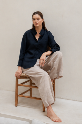 A woman sits barefoot on a wooden chair, wearing a dark navy button-up shirt and Olive Basics Jesse Wide-leg Pocket Pants in TENCEL™ Linen, set against a plain, light-colored background.