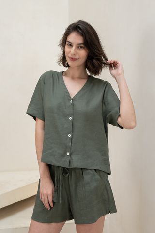 A woman stands indoors wearing the Olive Basics Bella Multi-way V-neck Shirt— a loose-fitting, short-sleeved, olive green button-up — paired with matching shorts, with one hand touching her hair.