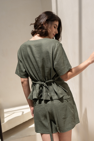 A woman stands indoors with her back to the camera, wearing the Olive Basics Bella Multi-way V-neck Shirt in olive green, paired with matching shorts.