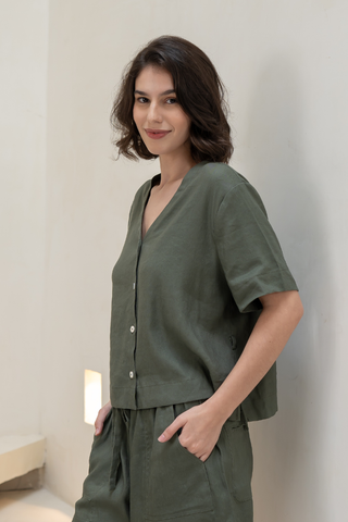 A woman with short brown hair wears the Olive Basics Bella Multi-way V-neck Shirt in olive green and matching pants, standing against a light-colored wall with one hand in her pocket.