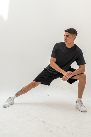 A person in a black shirt and Olive Basics Noah Multi-Pocket Stretch Shorts stretches their legs to the side while standing on a white floor against a plain background.