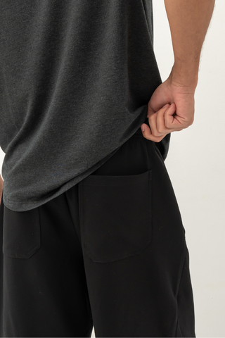 Wearing a dark shirt and Olive Basics Noah Multi-Pocket Stretch Shorts in black, a person stands with their hand gripping the hem of their shirt near their hip, facing away from the camera.