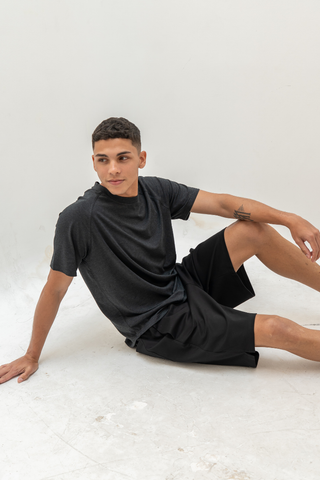 A person in a dark t-shirt and Olive Basics Noah Multi-Pocket Stretch Shorts in Powder Black sits on the floor against a plain white background, looking to the side with one arm propped behind them.
