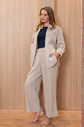 A person stands barefoot indoors against a light wood background, wearing Olive Basics’ Jesse Wide-leg Pocket Pants in TENCEL™ Linen (khaki) with a beige button-up shirt layered over a black top and hands in their pockets.