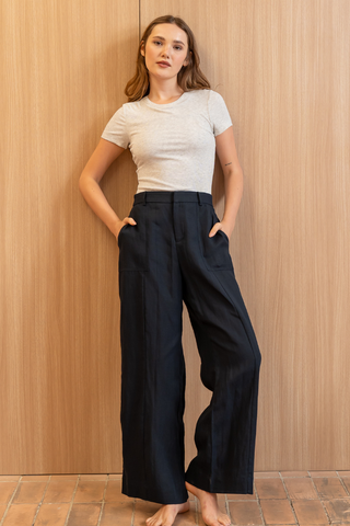 A woman stands barefoot indoors against a light wood wall, wearing a light gray short-sleeve t-shirt and the Olive Basics Jesse Wide-leg Pocket Pants in TENCEL™ Linen in Midnight Blue, with her hands in the pockets.