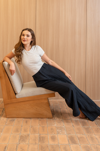 A woman wearing a grey t-shirt and the Olive Basics Jesse Wide-leg Pocket Pants in TENCEL™ Linen leans back on a wooden chair with light cushions, set against a wood-paneled wall and tile floor.