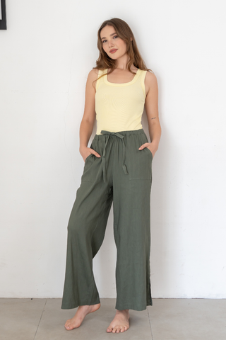 A woman stands barefoot against a white wall, wearing a yellow sleeveless top paired with Olive Basics’ Bella Multi-Pocket Drawstring Pants—loose green pants featuring multiple pockets, an adjustable waist, and her hands tucked casually in the pockets.
