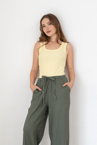A woman wearing the Olive Basics Anna Reversible Hi-Low Tank in Butter Yellow and green pants stands against a plain white wall with her hands in her pockets.