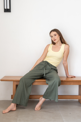 A woman sits barefoot on a wooden bench against a plain white wall, wearing a yellow tank top and Olive Basics’ Bella Multi-Pocket Drawstring Pants in green, featuring an adjustable waist.