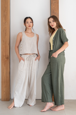 Two women stand indoors against a wood and white wall, wearing loose, casual outfits featuring sleeveless and short-sleeved tops paired with the Olive Basics Bella Multi-Pocket Drawstring Pants, crafted from a soft TENCEL linen blend. One wears the beige option and the other the green, both showcasing an adjustable waist design. Both women are barefoot.