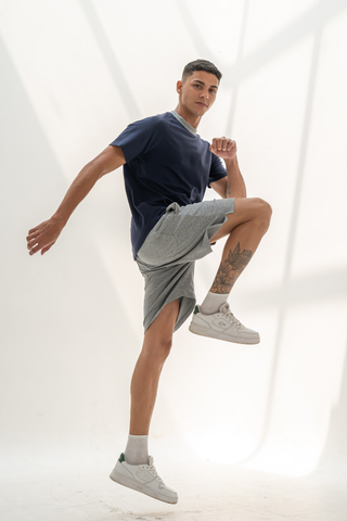 A young man in a navy t-shirt and Olive Basics Noah Multi-Pocket Stretch Shorts in heather grey poses mid-jump with one knee raised, balanced on one foot in a brightly lit studio.