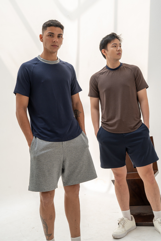 Two men stand side by side against a plain white background with soft natural lighting, wearing casual t-shirts paired with Olive Basics Noah Multi-Pocket Stretch Shorts, their hands in the shorts’ pockets.