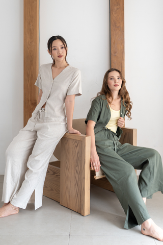 Two women wearing neutral-toned outfits pose indoors with a wooden chair, one seated and one standing, set against a minimal white and wood backdrop. One of them wears the Bella Multi-way V-neck Shirt by Olive Basics—a chic, nursing-friendly choice crafted from a linen blend.