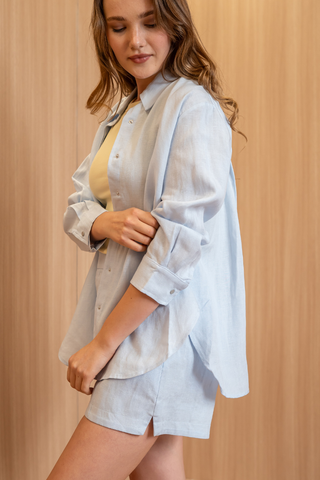 A woman wearing the Olive Basics Marie Everyday Shirt in TENCEL™ Linen and matching shorts stands against a light wooden background, looking down with one hand on her arm.