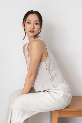 A woman with long dark hair sits on a wooden bench, wearing the Olive Basics Leigh-Anne Reversible Button Top in Heather Oatmeal paired with matching light-colored pants, against a plain white background.