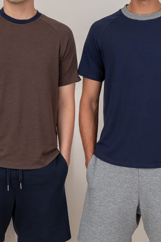 Two people stand side by side, each with hands in their pockets, wearing Olive Basics Noah Lightweight Multi-functional Tees—one styled in brown and navy, the other in navy and gray shorts. Their relaxed poses highlight the comfort of these lightweight TENCEL™ wool blend tees.