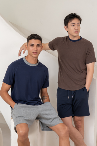 Two men wearing Olive Basics Noah Lightweight Multi-functional Tees and shorts pose indoors near a white staircase—one seated and one standing—with each looking in a different direction.